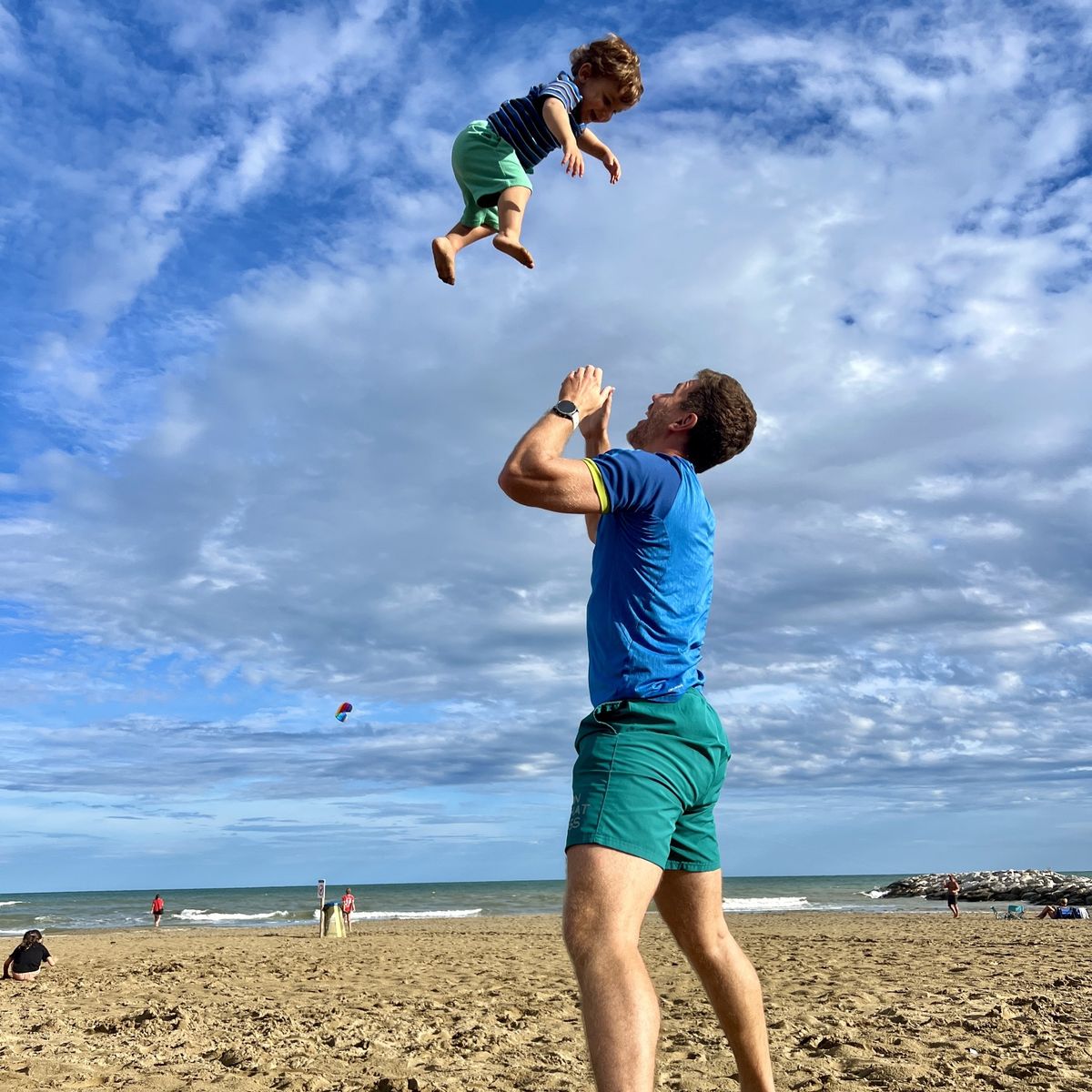 Sébastien Schmitt wirft am Strand lachend ein Kleinkind in die Luft, darüber blauer Himmel mit Wolken